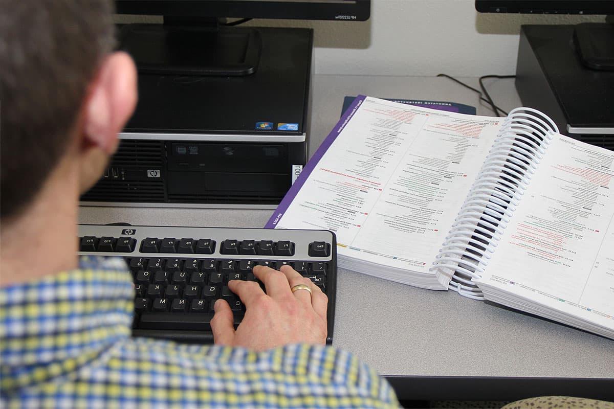 An individual sits at a computer, 在键盘上打字, with a book of medical coding information open on the desk next to them